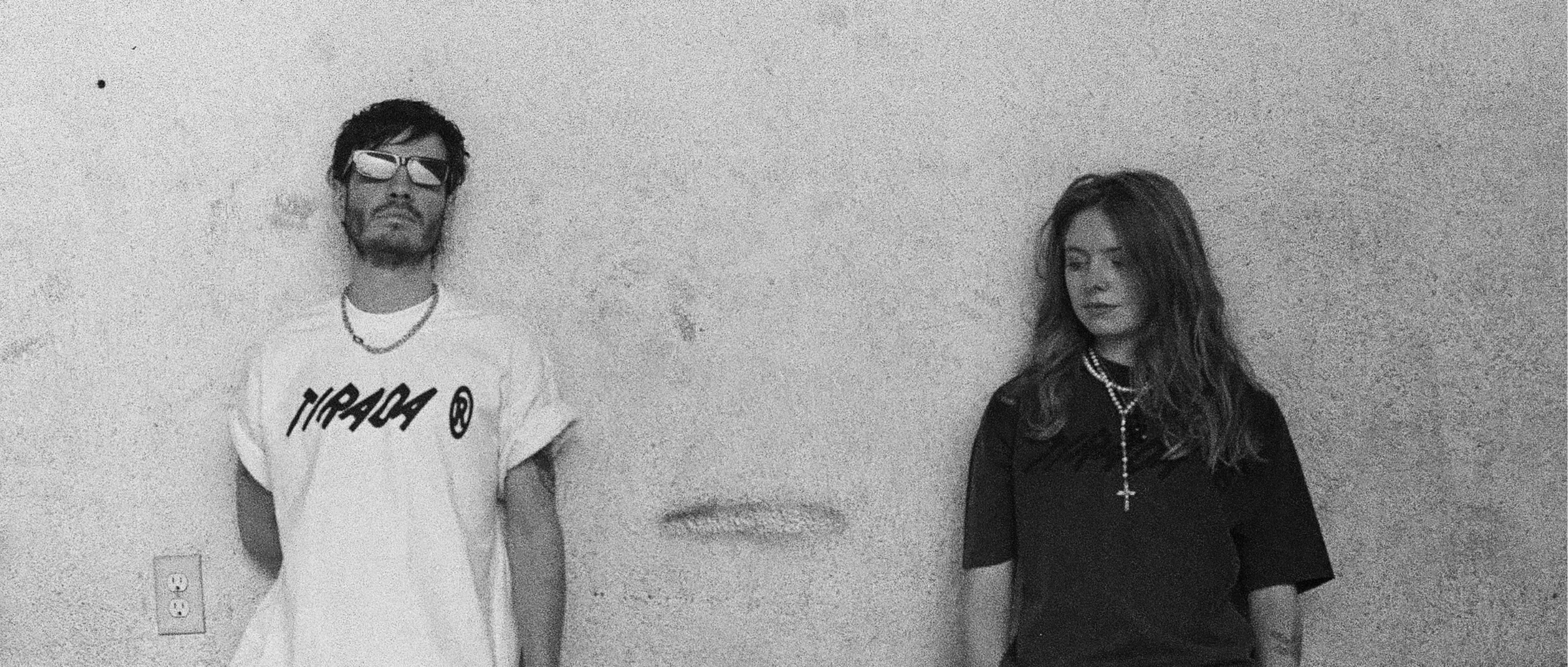 Chico con playera blanca de Tirada y chica con playera negra contra una pared de cemento, blanco y negro